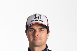O piloto Nelsinho Piquet, de boné branco e macacão de corrida azul-marinho com patrocínios, posa sorrindo em fundo branco