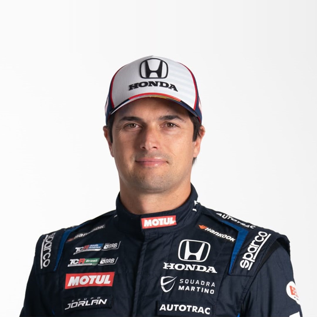 O piloto Nelsinho Piquet, de boné branco e macacão de corrida azul-marinho com patrocínios, posa sorrindo em fundo branco