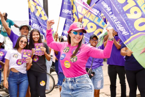 Danielle Garcia, de boné e óculos escuros rosa, ergue os braços em caminhada de campanha cercada de apoiadores com bandeiras roxas