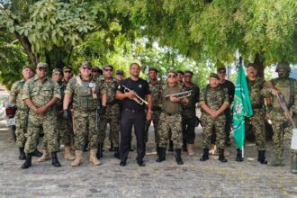 Grupo de veteranos do GVEB posa em fileira, em trajes militares, ao lado da bandeira do grupamento, sob árvores