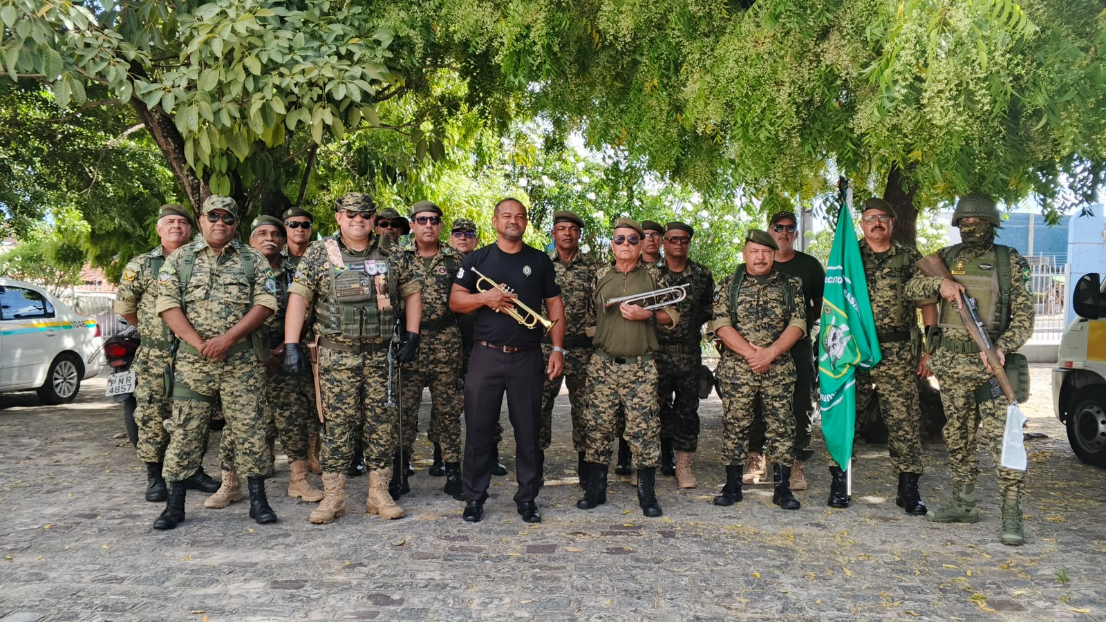 Grupo de veteranos do GVEB posa em fileira, em trajes militares, ao lado da bandeira do grupamento, sob árvores